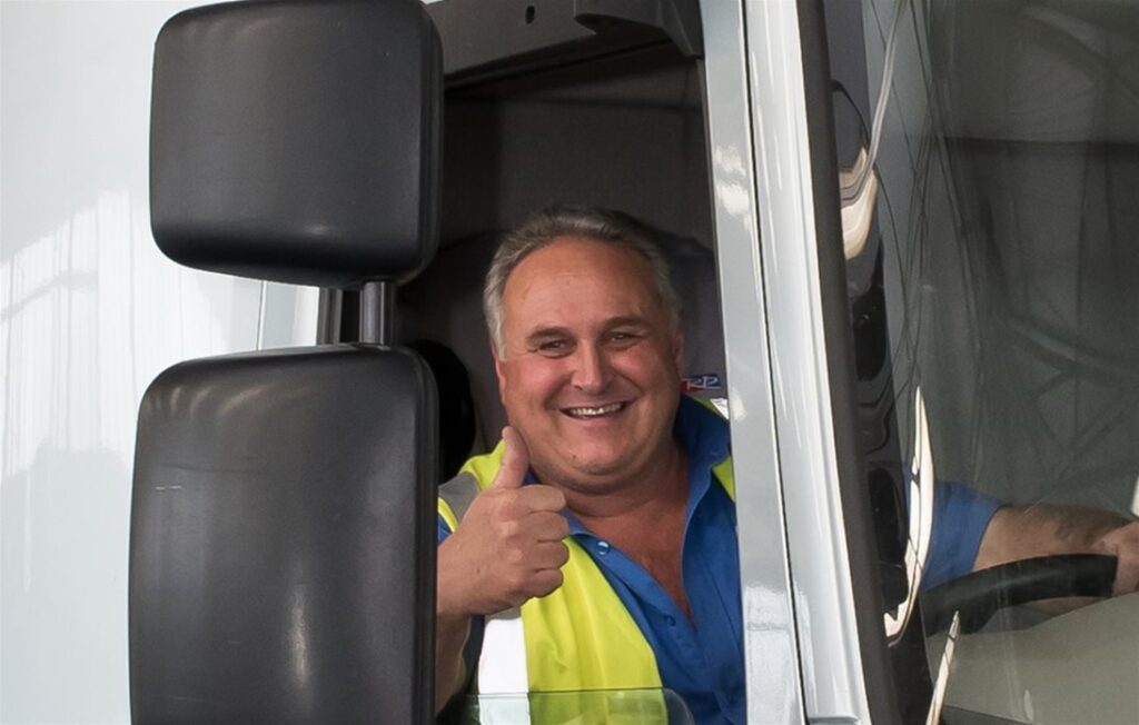 Smiling lorry driver giving a thumbs up from the cab of a white truck, wearing a high-visibility vest and blue shirt.