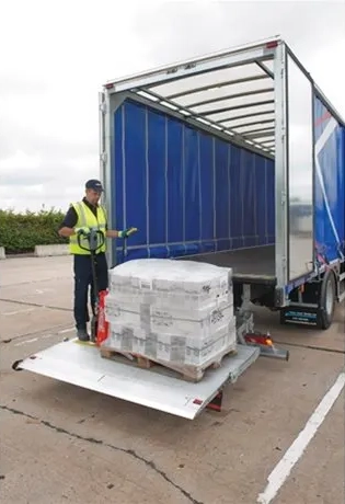 Delivery worker using a pallet truck to load a wrapped pallet of goods onto the tail lift of an open-sided Palletline lorry.