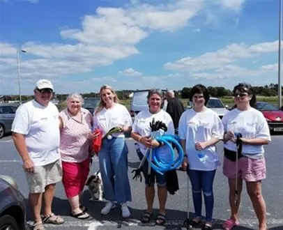 Group of six volunteers wearing white shirts and holding litter-picking tools, gathered in a sunny car park before a beach clean-up event, with cars and blue sky in the background.