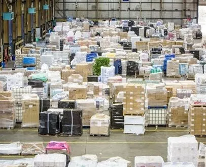 Large indoor distribution hub filled with assorted pallets of shrink-wrapped goods, boxes, and containers, each labeled and organised in designated zones